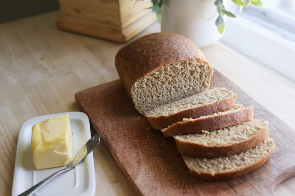 super fluffy sandwich bread with fresh milled flour sitting on a wooden cutting board with grain mill in background and butter nearby