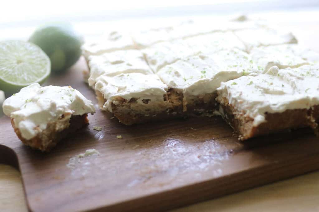 freshly milled key lime bars with whipped cream and lime zest on a cutting board that have been sliced