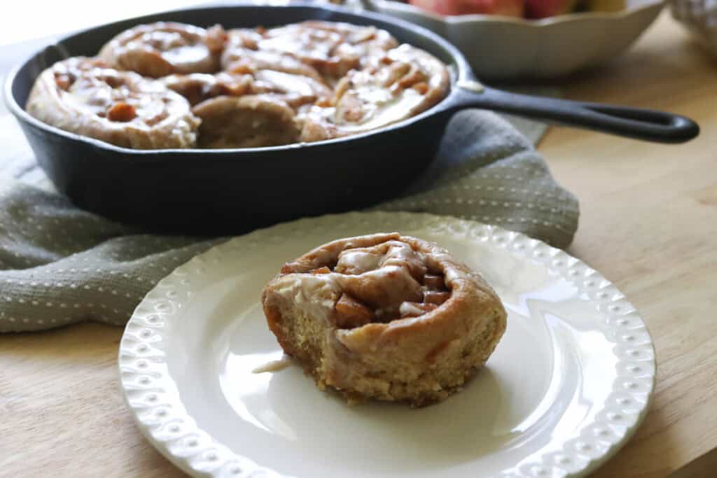 a freshly milled apple cinnamon roll on a white plate with a skillet of apple cinnamon rolls behind it