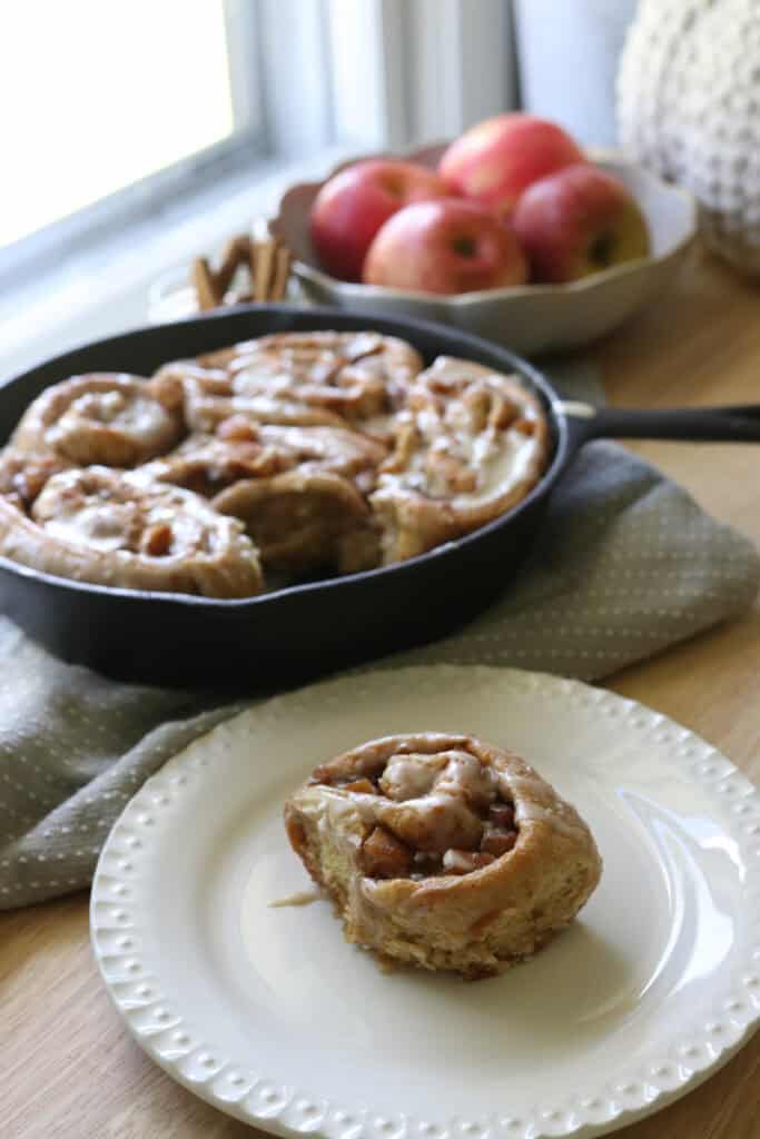 a  freshly milled apple cinnamon roll on a white plate with more freshly milled apple cinnamon rolls in a skillet