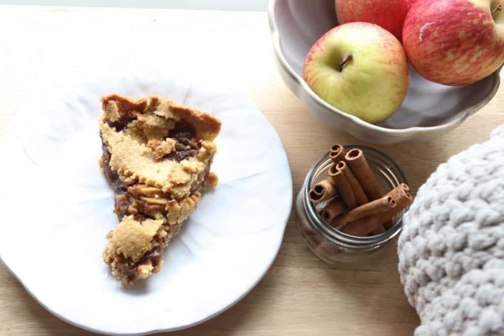 a slice of freshly milled classic apple pie on a plate with apples in a bowl and cinnamon sticks on a wooden background