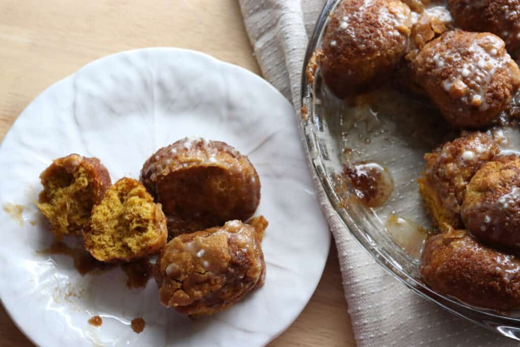 a scoop of fresh milled pumpkin monkey bread with glaze on top on a white plate with more monkey bread in a pie dish