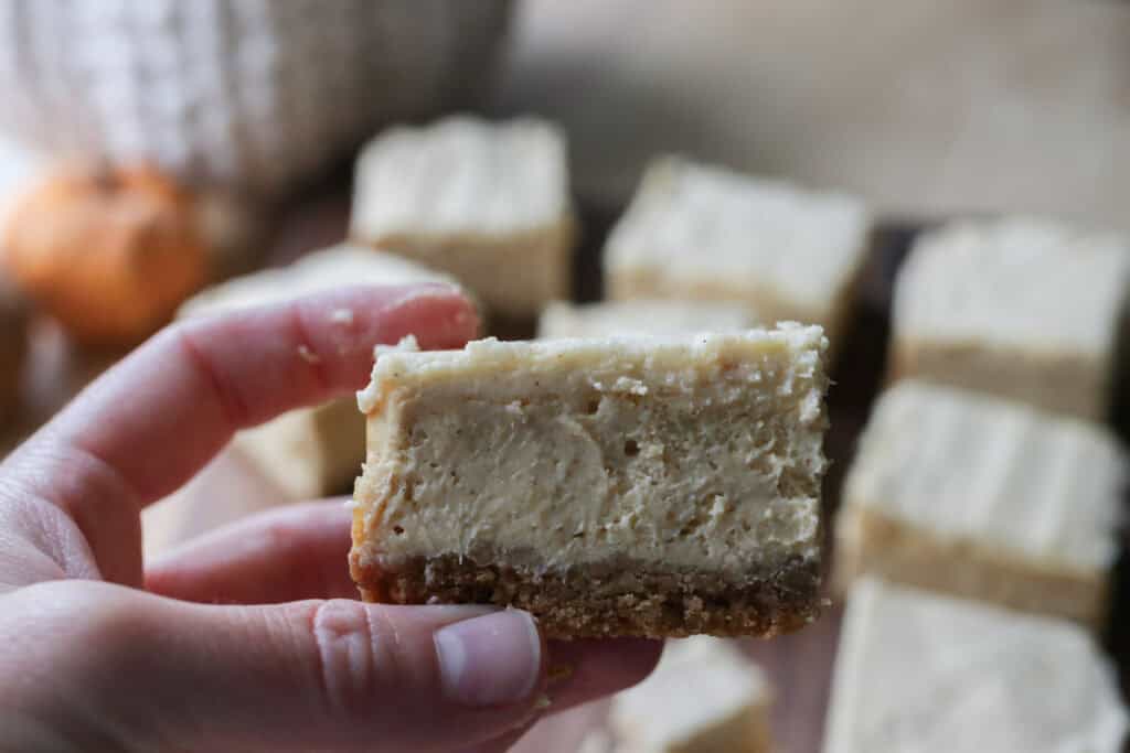 a pumpkin cheesecake bar with fresh milled crust being held by someone with more bars behind it