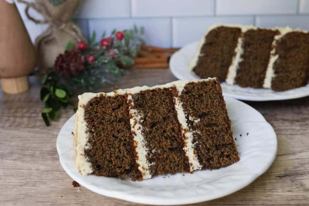 a slice of fresh milled gingerbread cake with cream cheese icing on a small white plate with another slice behind it