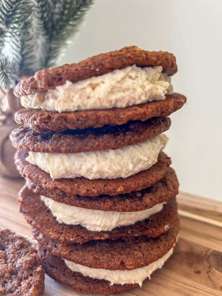 four Freshly Milled Chewy Gingerbread Oatmeal Cream Pies stacked together on a wooden cutting board with a tree in the background
