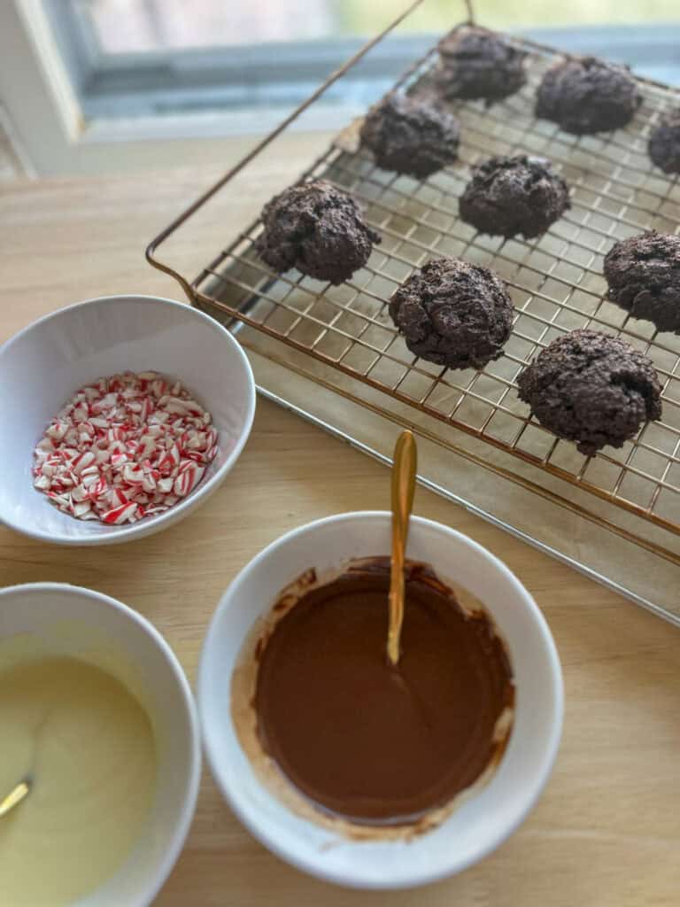 freshly milled double chocolate cookies on a wire rack with melted chocolate in white bowls and crushed candy canes in a bowl