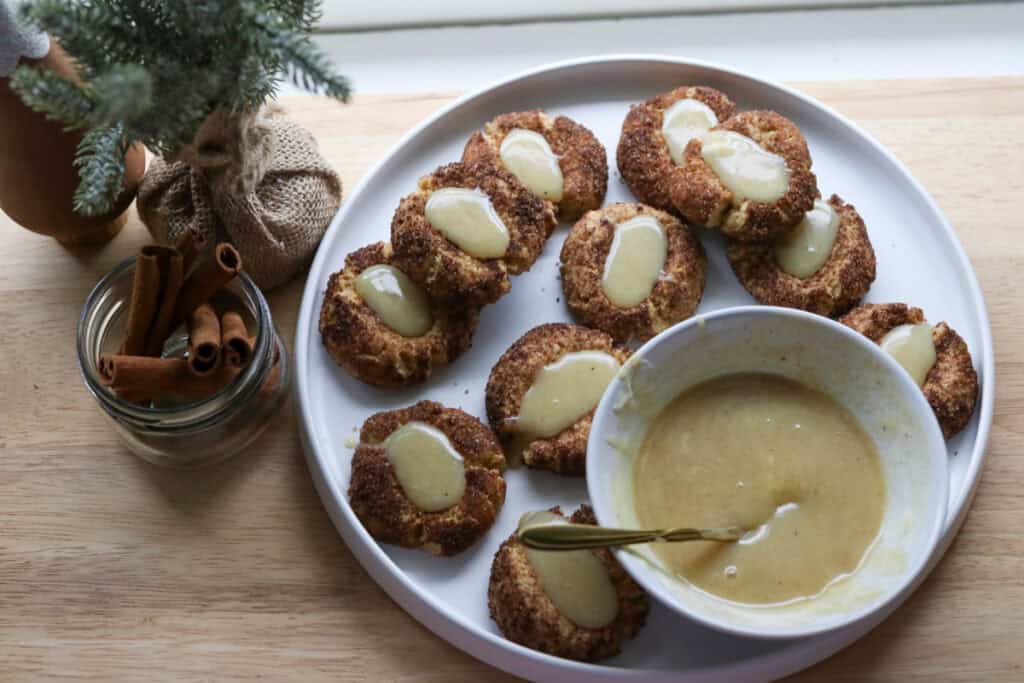 fresh milled eggnog snickerdoodle thumbprint cookies with a bowl of eggnog ganache on a white plate with cinnamon sticks