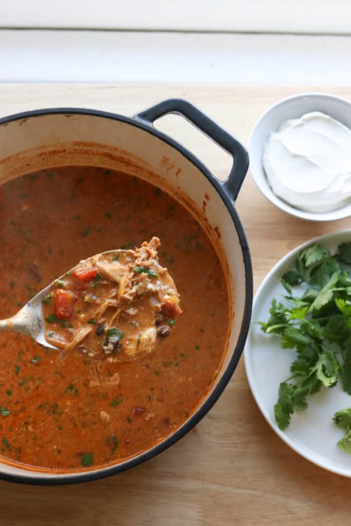a spoon of creamy chicken enchilada soup with a plate of cilantro and a bowl of sour cream