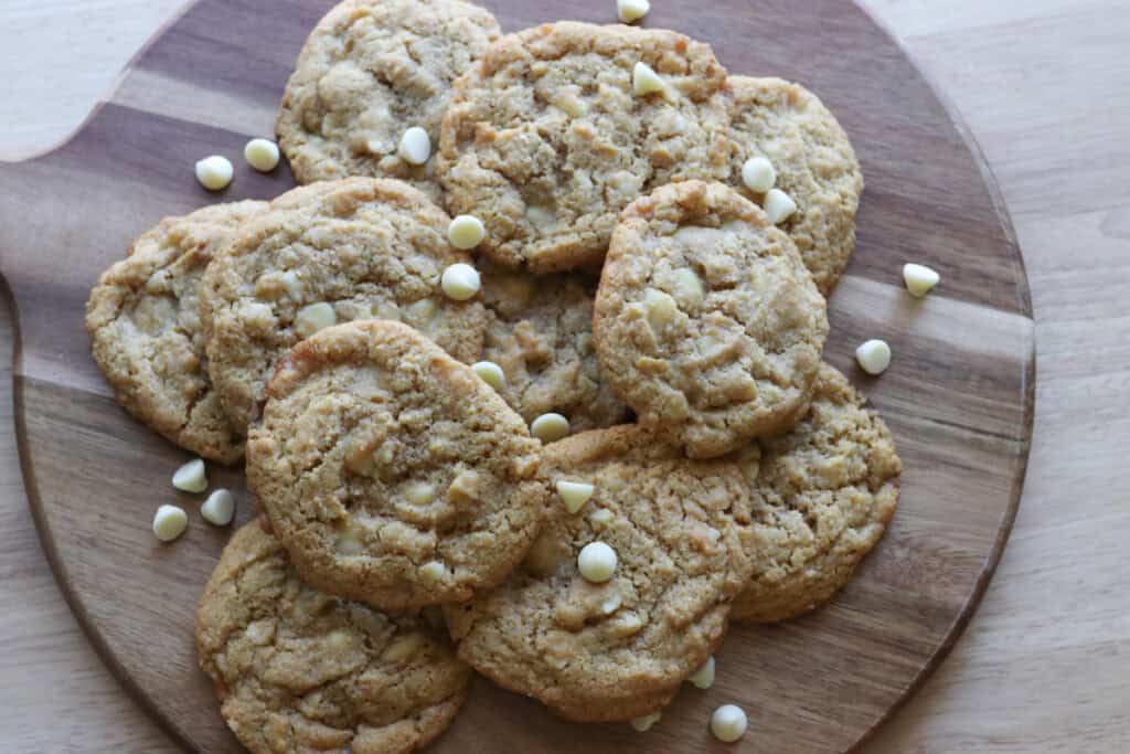 a bunch of Freshly Milled White Chocolate Macadamia Cookies on a wooden board with white chocolate chips on it