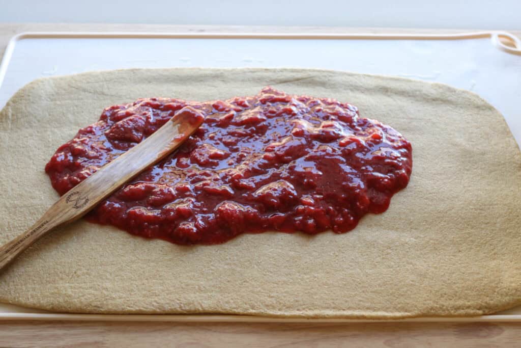 spreading strawberry filling overtop of Freshly Milled Valentine's Strawberry Cinnamon Roll dough