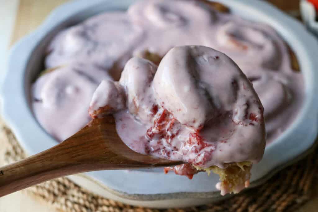 a spoon holding a Freshly Milled Valentine's Strawberry Cinnamon Roll with more cinnamon rolls behind it in a pan