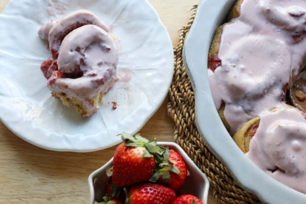 a Freshly Milled Valentine's Strawberry Cinnamon Roll on a white plate covered in frosting with strawberries in a bowl