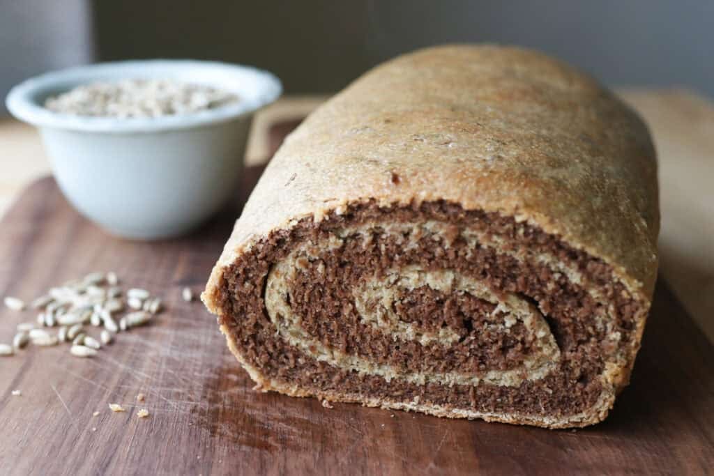 a loaf of freshly milled marbled rye bread that has been sliced with a bowl of rye wheat berries behind it