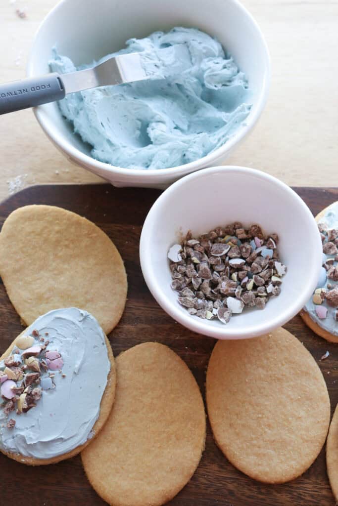 Fresh milled Easter egg sugar cookies on a board with blue frosting and crushed chocolate eggs in a bowl