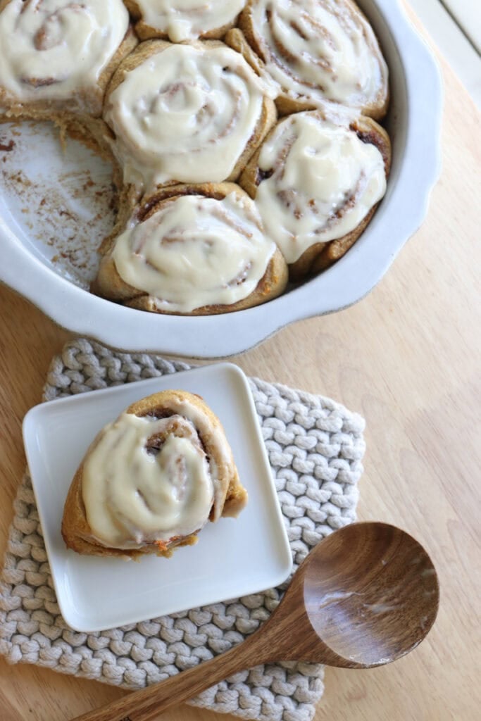 Fresh milled carrot cake cinnamon rolls in a dish with cream cheese icing on top and one on a plate