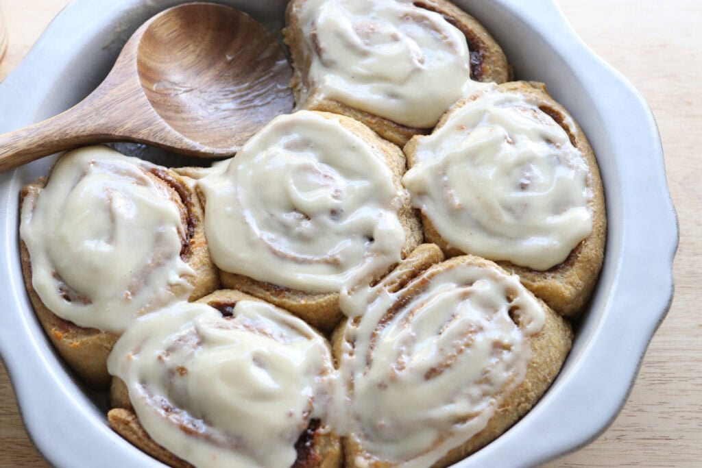 Fresh milled carrot cake cinnamon rolls topped with icing in a dish with a wooden spoon