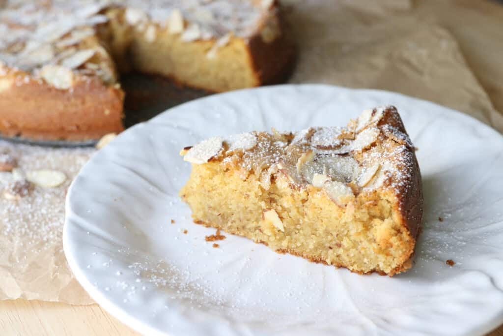 A slice of fresh milled almond croissant cake on a white plate with the cake behind it